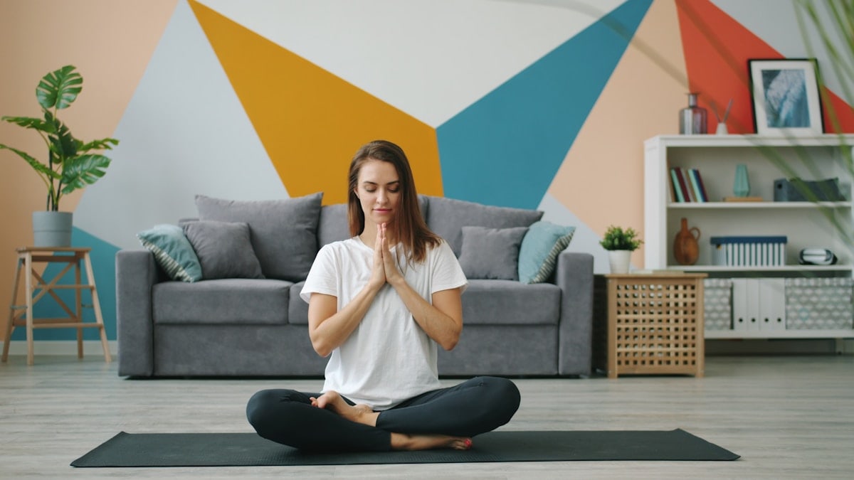 Attractive young woman in modern clothing is exercising at home in lotus position relaxing on yoga mat focused on practice. Healthcare and sports concept.